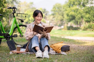 Güzel ve mutlu bir Asyalı kadın rahat bir şekilde yeşil bir parkta kitap okuyor, aydınlık bir günde..