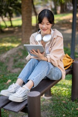 Olumlu, gülümseyen, günlük kıyafetler içindeki Asyalı bir kadın dijital tabletini parktaki bir bankta kullanıyor..