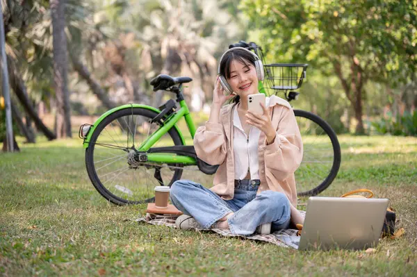 캐주얼 아시아 여자는 그녀의 스마트 공원에서 식히는 그녀의 헤드폰에 음악을 — 스톡 사진