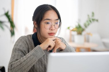 Zeki, düşünceli, Asyalı genç bir üniversite öğrencisi dizüstü bilgisayarından online bir raporu okumaya odaklanıyor, bir kafede uzaktan ödev yapıyor..