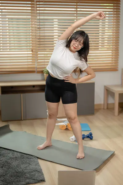 A strong, happy young Asian woman in sportswear is stretching her core muscles and arms on a yoga mat, working out or doing yoga at home.
