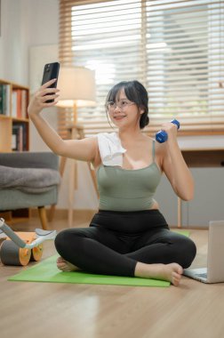 Spor giyim bölümünde genç ve güzel bir Asyalı kadın evde egzersiz yaparken akıllı telefonuyla selfie çekiyor. insanlar ve ev hayatı kavramları