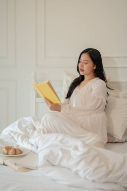 Uzun pijamalı güzel, sakin bir Asyalı kadın sabah yatağında kitap okuyor. ev hayatı ve boş zaman kavramları