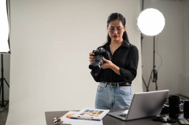 Profesyonel, çekici bir Asyalı kadın fotoğrafçı DSLR kamerasını bir fotoshop için ayarlıyor. Profesyonel bir stüdyoda ışıklandırma ekipmanlarıyla çalışıyor..