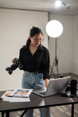 Profesyonel, çekici bir Asyalı kadın fotoğrafçı dizüstü bilgisayarındaki görüntüleri kontrol ediyor, profesyonel ışıklandırma ekipmanlarıyla bir stüdyoda çalışıyor..
