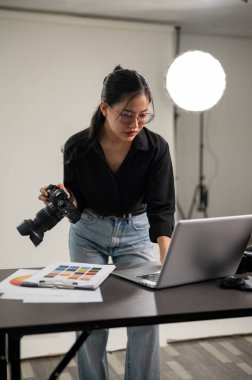 Profesyonel, çekici bir Asyalı kadın fotoğrafçı dizüstü bilgisayarındaki görüntüleri kontrol ediyor, profesyonel ışıklandırma ekipmanlarıyla bir stüdyoda çalışıyor..