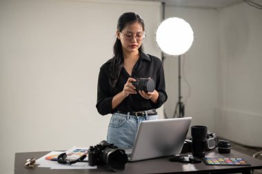 Profesyonel, çekici bir Asyalı kadın fotoğrafçı kamerasını foto çekimine hazırlıyor, profesyonel ışıklandırma ekipmanlarıyla bir stüdyoda çalışıyor..