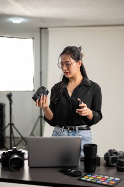 Profesyonel, çekici bir Asyalı kadın fotoğrafçı kamerasını foto çekimine hazırlıyor, profesyonel ışıklandırma ekipmanlarıyla bir stüdyoda çalışıyor..