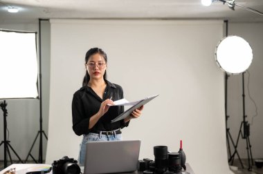 Profesyonel, çekici bir Asyalı kadın fotoğrafçı, fotoshoot 'un detaylarını kontrol etmeye odaklanıyor. Stüdyosunda profesyonel ışıklandırma ekipmanlarıyla çalışıyor..