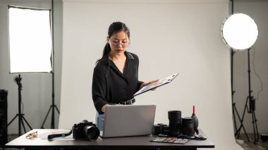 Profesyonel, çekici bir Asyalı kadın fotoğrafçı, fotoshoot 'un detaylarını kontrol etmeye odaklanıyor. Stüdyosunda profesyonel ışıklandırma ekipmanlarıyla çalışıyor..