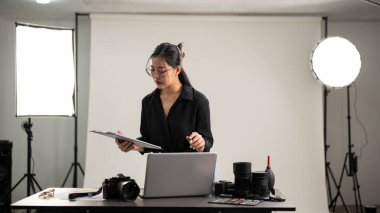 Profesyonel, çekici bir Asyalı kadın fotoğrafçı, fotoshoot 'un detaylarını kontrol etmeye odaklanıyor. Stüdyosunda profesyonel ışıklandırma ekipmanlarıyla çalışıyor..