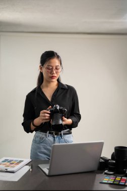 DSLR kamerasıyla siyah gömlekli zarif, profesyonel bir Asyalı kadın fotoğrafçı stüdyosunda duruyor..