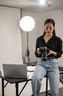 Profesyonel bir Asyalı kadın fotoğrafçı DSLR kamerasını ayarlamaya odaklanıyor, ışıklandırma ekipmanlarıyla bir fotoshoot stüdyosunda çalışıyor..