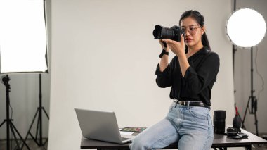 Profesyonel, kendine güvenen bir Asyalı kadın fotoğrafçı fotoğraf çekiyor, yüzünün yanında bir kamera tutuyor, kamerasını ayarlıyor, profesyonel ışıklandırma ekipmanlarıyla bir stüdyoda çalışıyor..