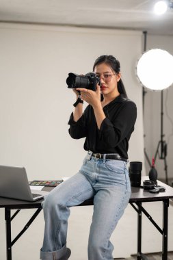 Profesyonel, kendine güvenen bir Asyalı kadın fotoğrafçı fotoğraf çekiyor, yüzünün yanında bir kamera tutuyor, kamerasını ayarlıyor, profesyonel ışıklandırma ekipmanlarıyla bir stüdyoda çalışıyor..