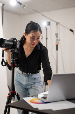 Havalı, profesyonel bir Asyalı kadın fotoğrafçı dizüstü bilgisayarındaki görüntüleri kontrol ediyor. Stüdyoda profesyonel fotoğraf ekipmanlarıyla çalışıyor..