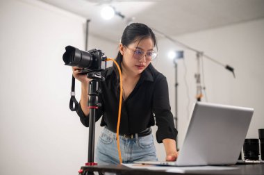 Havalı, profesyonel bir Asyalı kadın fotoğrafçı dizüstü bilgisayarındaki görüntüleri kontrol ediyor. Stüdyoda profesyonel fotoğraf ekipmanlarıyla çalışıyor..