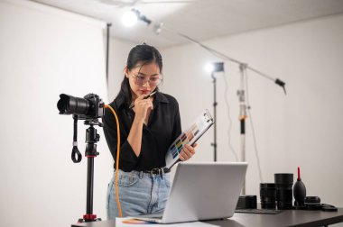 Çekim stüdyosunda çalışan havalı, çekici bir Asyalı kadın fotoğrafçı dizüstü bilgisayarındaki görüntüleri kontrol ediyor. Profesyonel fotoğraf ışıklandırma ekipmanlarının olduğu bir stüdyo.