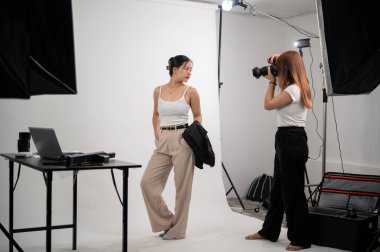 Profesyonel bir kadın fotoğrafçı, sıradan giyinmiş çekici bir Asyalı kadının fotoğraflarını çekiyor. Profesyonel ışıklandırma ekipmanlarıyla bir stüdyoda moda fotoğrafçılığı yapıyor..