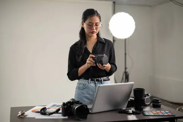 A professional, attractive Asian female photographer is preparing her camera for a photoshoot, working in a studio with professional lighting equipment.