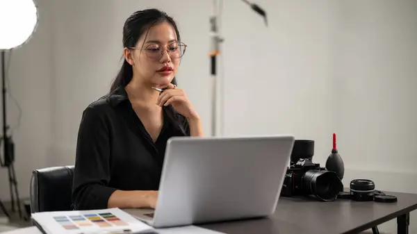 A confident, professional Asian female photographer is working on her laptop computer, checking images, working in a photoshoot studio with professional lighting equipment.