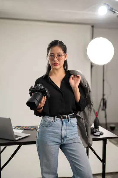 A confident, professional Asian female photographer in a black shirt is standing in her photoshoot studio with a DSLR camera in her hand.
