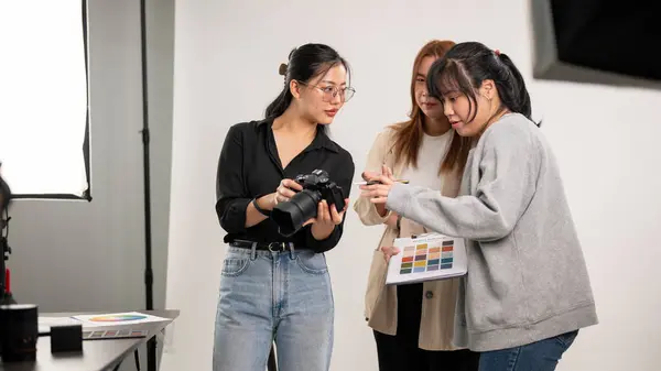 A cool, professional Asian female photographer is talking and showing images on the camera to her team, working in the fashion shooting studio, and briefing the concept with an art director.