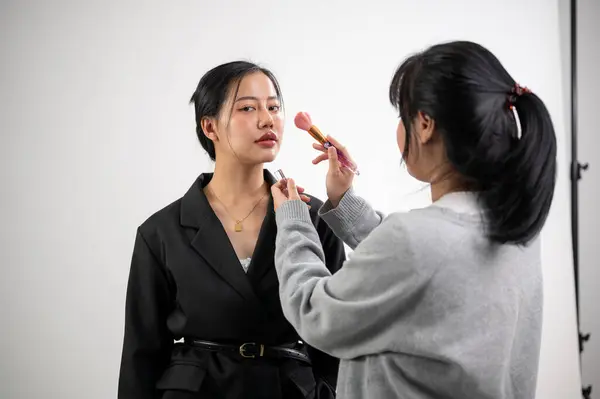 A beautiful young Asian female model is having makeup applied by a female makeup artist on a set, preparing for a fashion magazine photoshoot in the studio with professional lighting equipment.