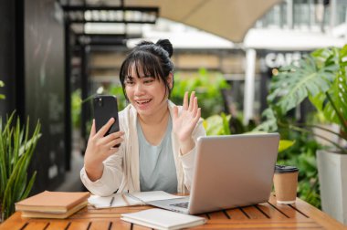 Sıradan giyinmiş genç, pozitif Asyalı bir kadın açık havada uzaktan çalışan biriyle görüntülü konuşma yapıyor. İnsanlar ve kablosuz teknoloji kavramları