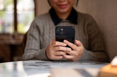 Kapalı bir masada akıllı telefonunu kullanan Asyalı bir kadının görüntüsü, mesajları kontrol eden, sohbet eden, sosyal medyada kaydıran ve internetten mal sipariş eden. İnsanlar ve kablosuz teknoloji kavramları
