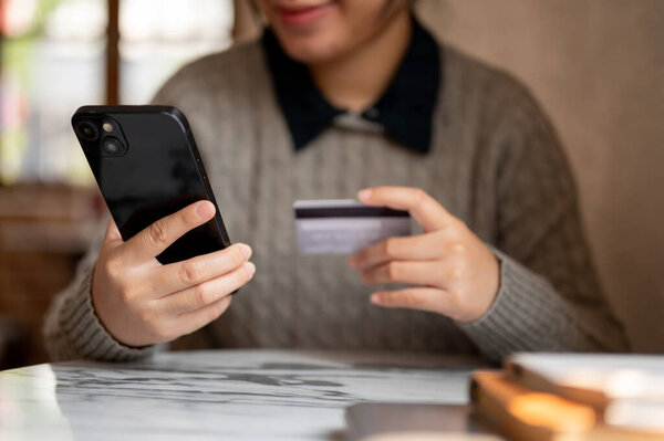 A cropped shot of an Asian woman holding her smartphone and a credit card, sitting at a table indoors, shopping online, registering a credit card account on a shopping website, or transferring money.