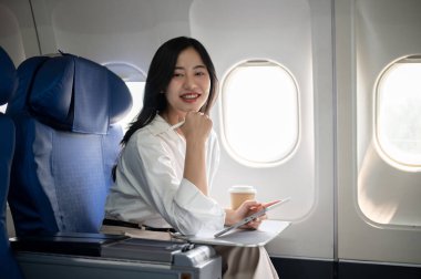 Çekici, kendine güvenen Asyalı bir iş kadını, bir iş seyahati için uçakta birinci sınıf bir uçağın cam kenarında otururken kameraya gülümsüyor. iş insanları ve ulaşım