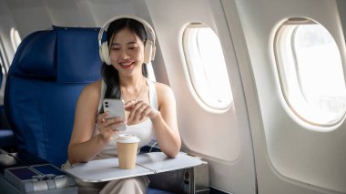 Sıradan giyinmiş çekici, gülümseyen genç bir Asyalı kadın uçuş sırasında akıllı telefonunu kullanıyor ve kulaklıkla müzik dinliyor, uçakla seyahat ediyor. Yaşam tarzı, ulaşım, turizm