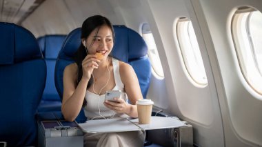 Çekici bir Asyalı bayan yolcu uçuş sırasında aperatiflerin ve kulaklıklarla müzik dinlemenin keyfini çıkarıyor, yaz seyahati için uçakla seyahat ediyor. insanlar ve ulaşım kavramları