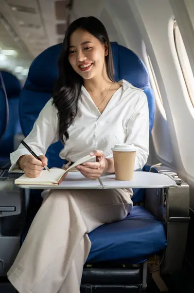 An attractive Asian businesswoman is taking notes in her notebook while flying to another city for a business meeting. A businesswoman is on the plane, sitting at the window seat in business class.