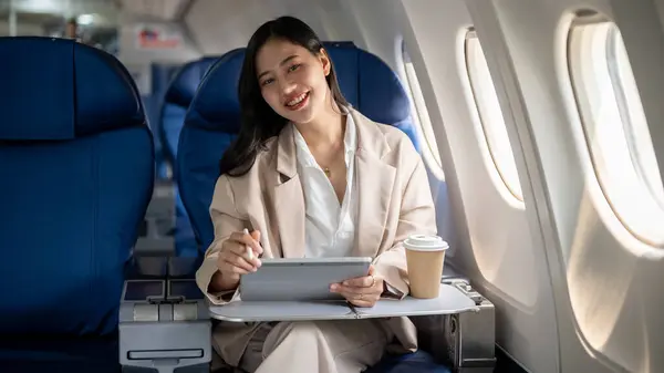 A confident, attractive Asian businesswoman is sitting at a window seat on a plane with her digital tablet, smiling at the camera, flying for a business trip. businesspeople and transportation