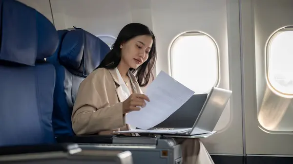 An attractive, confident, hard-working Asian businesswoman is working on her laptop computer, reading documents, and preparing her report during the flight for her business trip.