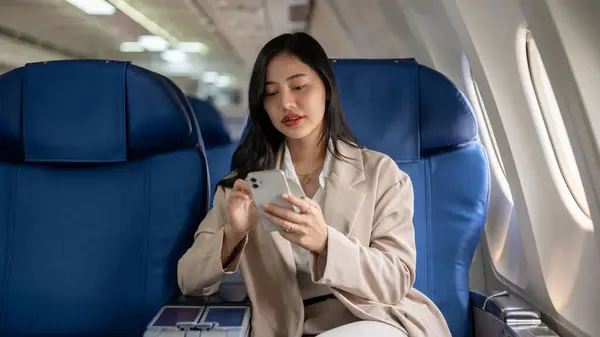 An attractive Asian businesswoman in a business suit is checking messages or turn on the airplane mode on her smartphone during the flight or before takeoff, flying to a business meeting.