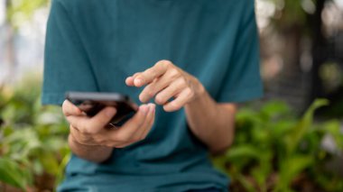 Sıradan giyinmiş bir adamın açık havada akıllı telefonunu kullanırken, parkta bir bankta otururken çekilmiş görüntüsü. Sosyal medya, ağ, sohbet, mobil uygulama