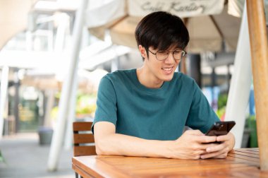 Gözlüklü gülümseyen genç Asyalı bir adam, dışarıdaki ahşap bir masada oturuyor, akıllı telefonuna bakıyor, biriyle sohbet ediyor ya da sosyal medyasına kayıyor. İnsanlar ve kablosuz teknoloji kavramları