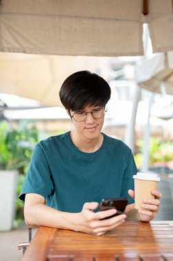 Gözlüklü genç bir Asyalı, elinde akıllı telefon ve tek kullanımlık kahve fincanı olan bir kafenin ahşap masasında oturuyor. İnsanlar ve modern şehir hayatı kavramları