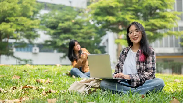 在一个阳光明媚的日子里 一位笑着的年轻亚洲女学生正坐在校园公园的草地上 她用笔记本电脑学习 另一位学生则是她的背景 大学生活概念 — 图库照片