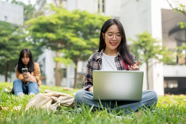 一位年轻的亚洲女大学生正坐在校园公园的草地上 在她的笔记本电脑上和她的朋友们举行电视电话会议 无线科技和教育概念 — 图库照片