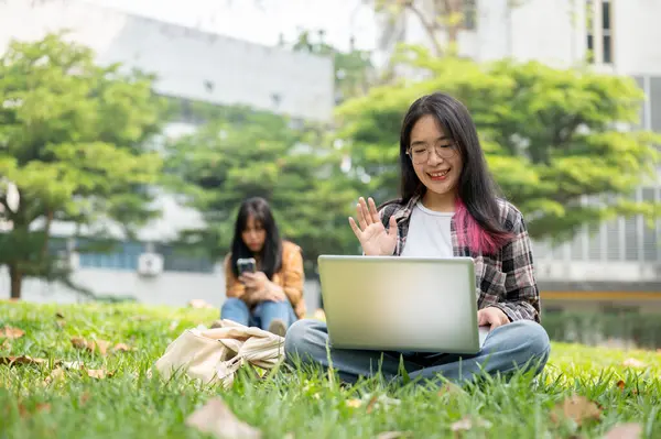 一位年轻的亚洲女大学生正坐在校园公园的草地上 在她的笔记本电脑上和她的朋友们举行电视电话会议 无线科技和教育概念 — 图库照片