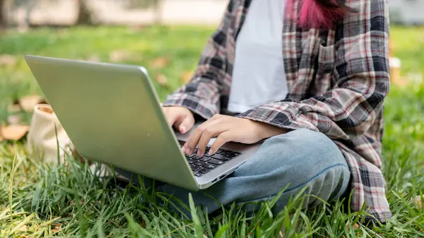 A cropped image of a female college student in casual wear is using her laptop computer while sitting on the grass in a park on a sunny day. wireless technology concept