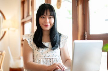 Güzel bir Asyalı kadın bir kafede dizüstü bilgisayarıyla oturuyor uzaktan çalışıyor, kameraya gülümsüyor. insanlar ve teknoloji kavramları