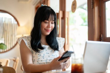 Günlük kıyafetlerle mutlu bir Asyalı kadın güzel, rahat bir kahve dükkanında oturuyor, akıllı telefonunu kullanıyor, mesajları kontrol ediyor ya da sohbete cevap veriyor, bir kafede uzaktan çalışıyor..