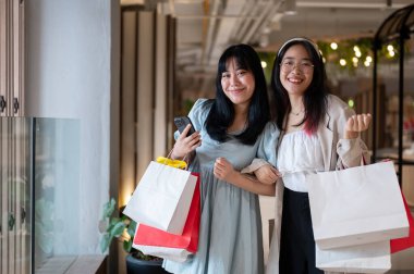 Alışveriş merkezinde alışveriş torbaları tutan, kameraya gülümseyen, birlikte alışveriş yapan iki neşeli Asyalı kadın. Alışveriş ve yaşam tarzı kavramları