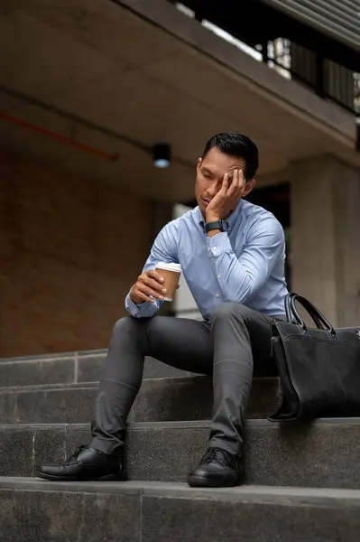 A stressed Asian millennial businessman, appearing tired and unhappy, sits on the steps outside a building, holding a coffee cup and covering his face, feelings of failure and exhaustion.