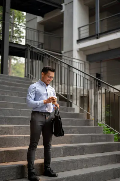 A handsome, confident Asian millennial businessman stands on the stairs outside of the building, holding a coffee cup and his briefcase. business people concept
